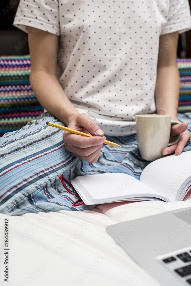 A woman is sitting in bed on a blanket in her hands holding a cup of hot drink and preparing to make a note in a notebook with a pencil, next to her laptop. A woman is sitting in bed on a blanket in her hands holding a cup of hot drink and preparing to make a note in a notebook with a pencil, next to her laptop.