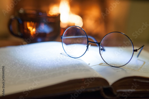 An open ancient book, antique glasses and a cup of hot drink in front of the fireplace. Evening fairy tales. Fantasy.