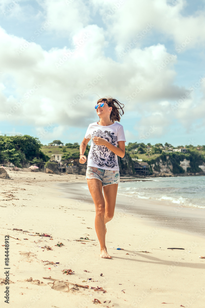 Sporty and healthy young woman in sunglasses running on the tropical beach during sunset. Bali island, Indonesia.