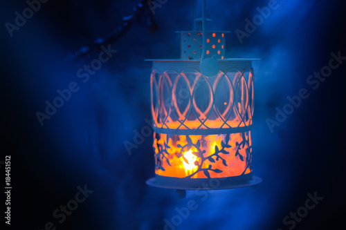 Decorative Christmas lantern with burning candle hanging on snow-covered fir- tree branch in a winter park. New year festive card, poster, postcard design.