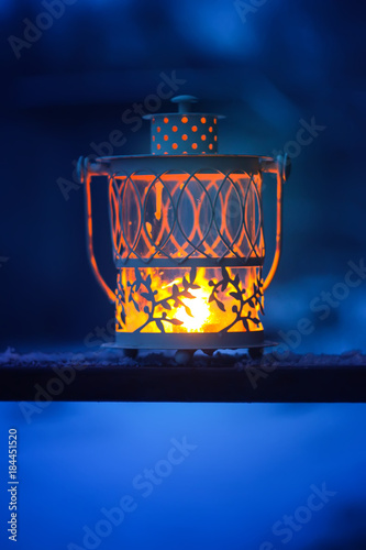 Decorative Christmas lantern with burning candle hanging on snow-covered fir- tree branch in a winter park. New year festive card, poster, postcard design.
