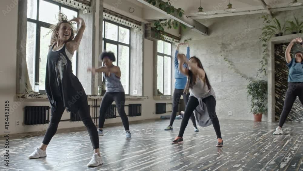 PAN of female dance instructor showing warming up exercise to multiethnic group of young women in class