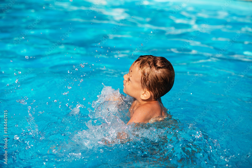 Happy kid playing in blue water of swimming pool. Little boy learning to swim. Summer vacations concept. Cute boy swimming in pool water. Child splashing in swimming pool
