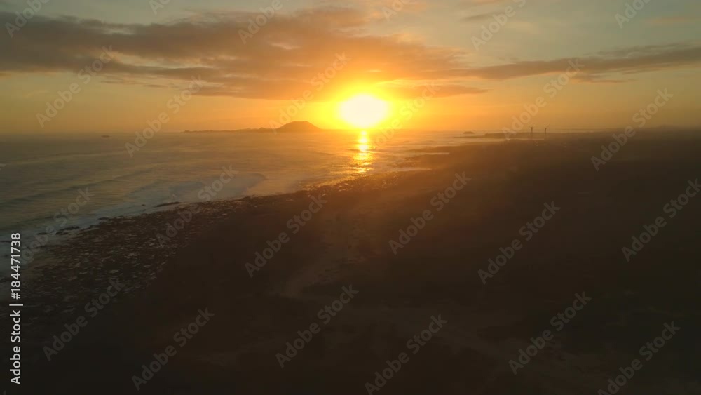 AERIAL: Flying over breathtaking beaches of Corralejo at sunset. Picturesque view of the setting sun behind a volcano as foaming waves reach rocky coast. Wind farm turbines in the distance. AERIAL: Flying over breathtaking beaches of Corralejo at sunset. Picturesque view of the setting sun behind a volcano as foaming waves reach rocky coast. Wind farm turbines in the distance.