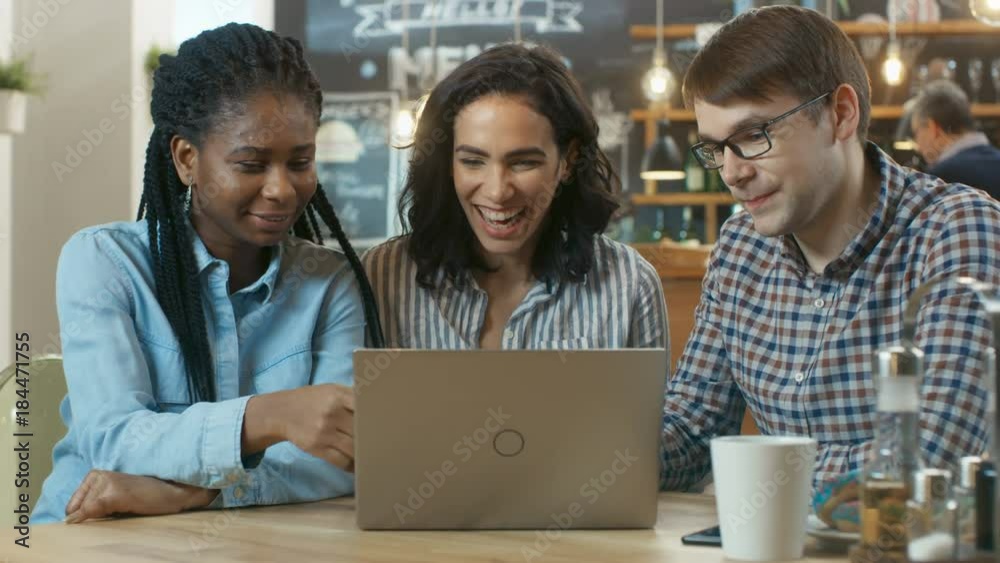 Three Young People Have Fun in the Coffee Shop, They Watch Something on a Laptop and Talk. In the Background Stylish Cafe Establishment. Shot on RED EPIC-W 8K Helium Cinema Camera. Three Young People Have Fun in the Coffee Shop, They Watch Something on a Laptop and Talk. In the Background Stylish Cafe Establishment. Shot on RED EPIC-W 8K Helium Cinema Camera.