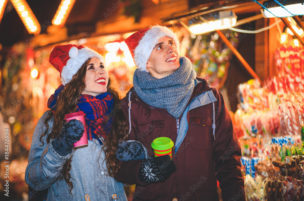 Happy couple in warm clothes with hot drinks enjoying Christmas market at evening