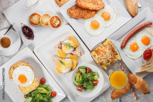 Breakfast table with egg Benedict, fried eggs and sausages, toasts with fried eggs and cheese, curd fritters. Fresh orange juice and coffee with bread rolls and croissants. Top view, flat lay