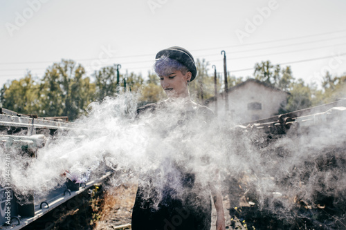 .Young modern tattoed woman dressed in black at an abandoned train station, playing with colorful smoke between train tracks. Lifestyle portrait