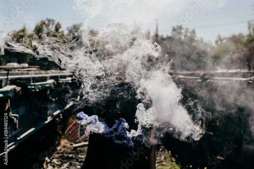 .Young modern tattoed woman dressed in black at an abandoned train station  playing with colorful smoke between train tracks. Lifestyle portrait