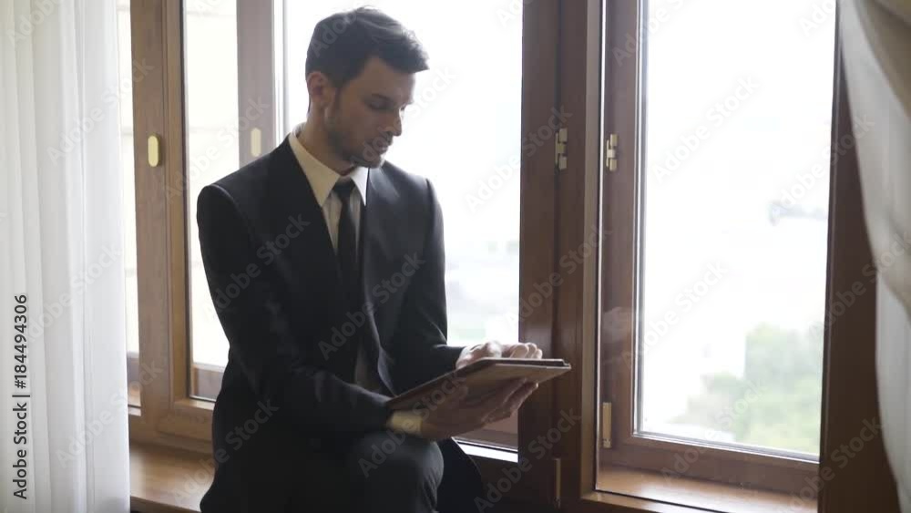 Young businessman is sitting on a windowsill and working with his tablet computer. Handheld real time establishing shot