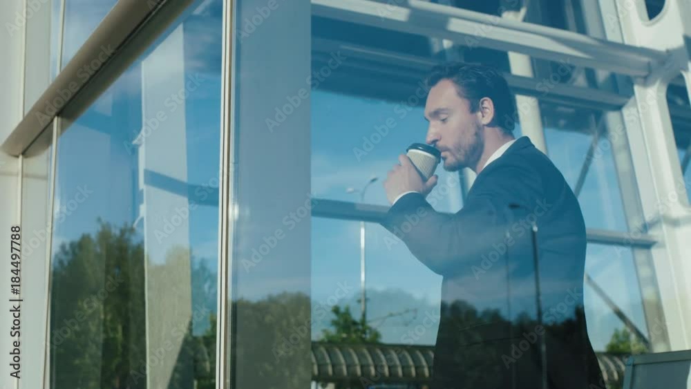 handsome businessman in a tie and suit drinking coffee to go and looking woth a smile out of the window of his office and than going away. Sunny day. Indoor