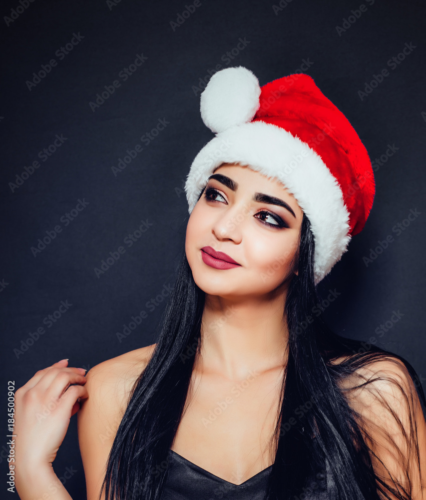 A beautiful woman with black straight hair and a Santa Claus hat on a dark background looks away. Close-up. Space for text A beautiful woman with black straight hair and a Santa Claus hat on a dark background looks away. Close-up. Space for text