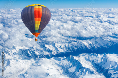 Aerial view from colorful hot air balloon flying over mountains with snow and clouds.