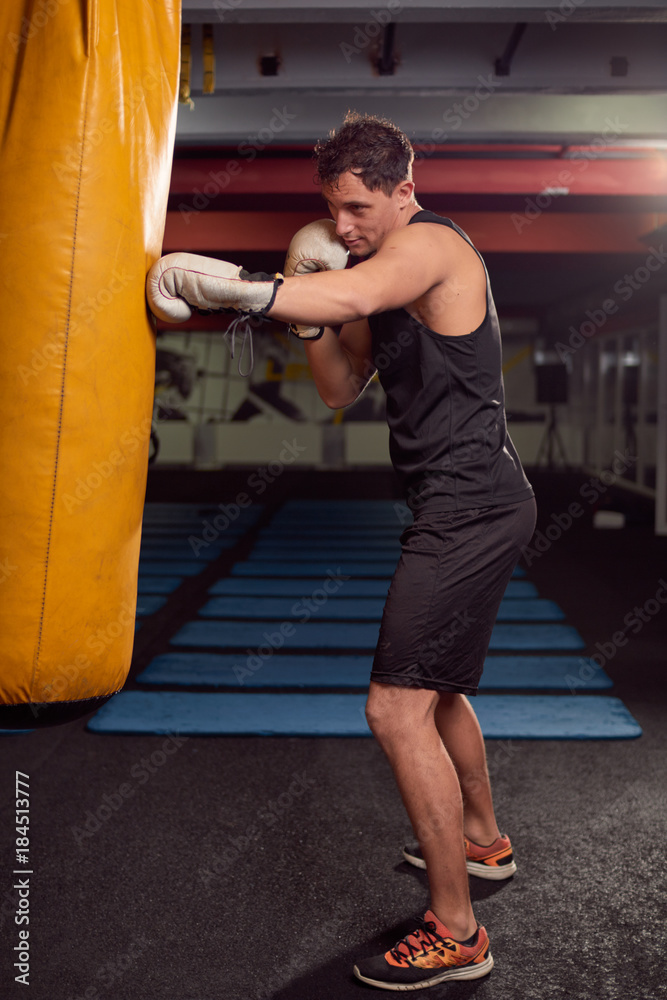 one young man, boxer hand hitting punching bag, practicing indoors gym room, wearing boxing gloves, full lenght body shot.