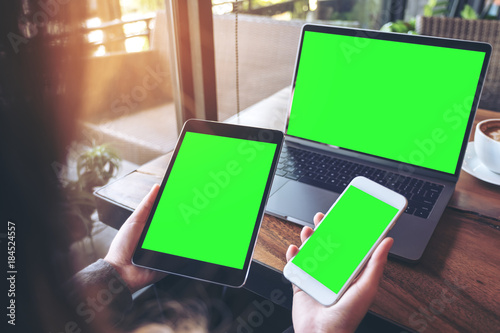 Mockup image of a businesswoman holding white mobile phone , black tablet and laptop with blank green screen on vintage wooden table with coffee cup in cafe