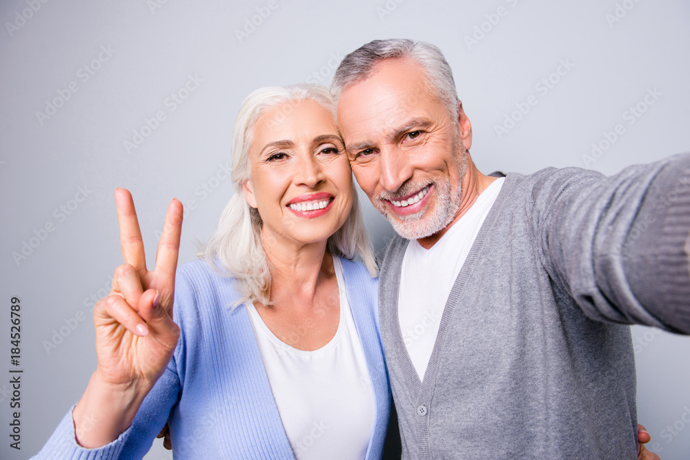 Happy sweet memories! Close up photo of senior couple showing two fingers and taking a selfie, isolated on grey background Happy sweet memories! Close up photo of senior couple showing two fingers and taking a selfie, isolated on grey background