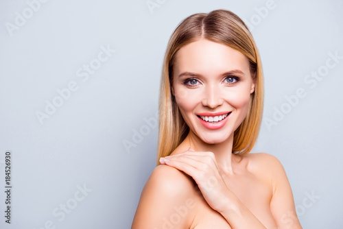 Close up portrait of beautiful lovely and smiling young woman with ideal skin and hair. She is touching her shoulder and enjoying the affect after cosmetological procedure. Isolated on grey background