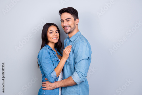 Portrait of caucasion lovely couple - man with bristle embrace his charming, cute woman and they both looking at camera while standing over grey background