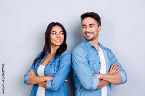 Portrait of caucasion cute hispanic couple - man with bristle and pretty, cute woman with crossed hands standing half turned and looking to each other over grey background
