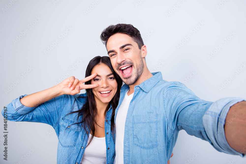 Sweet, caucasian, cheerful lovely couple in casual outfit - man making self portrait, woman showing peace symbol near her eye, standing over grey background Sweet, caucasian, cheerful lovely couple in casual outfit - man making self portrait, woman showing peace symbol near her eye, standing over grey background