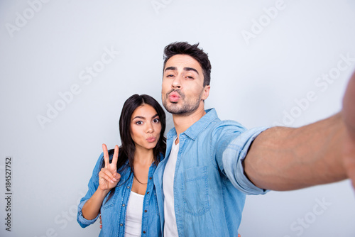 Sweet, attractive, hispanic, caucasian couple - man making self portrait, woman showing peace symbol, they sending kiss to the camera, standing over grey background