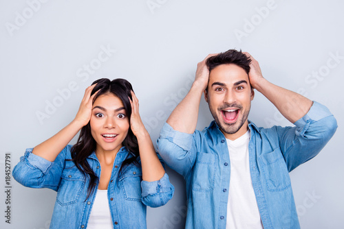  Portrait of two young surprised lovers  with wide opened eyes holding heads in jeans shirts standing over grey background