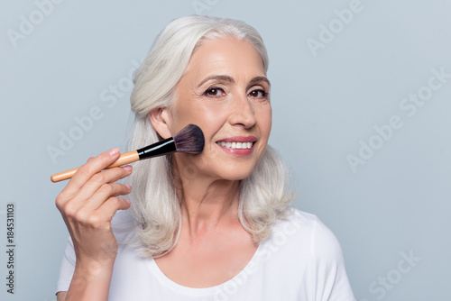 Middle aged, attractive, caucasian, nice woman applying makeup on her face, using powder, rouge, holding tassel in hand, making cheekbones over grey background