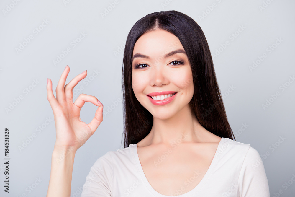 Close up portrait of happy, pretty, confident, stylish, smiling girl in white t-shirt showing ok sign standing on grey background Close up portrait of happy, pretty, confident, stylish, smiling girl in white t-shirt showing ok sign standing on grey background