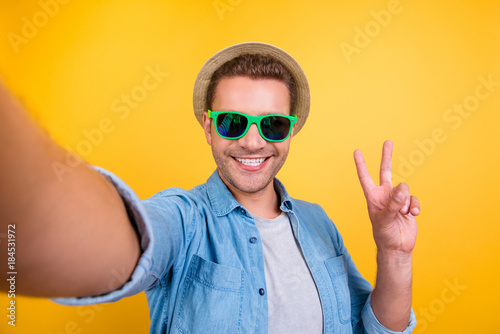 Self portrait of cheerful, bearded blogger in summer glasses, jeans shirt shooting selfie, showing peace symbol with two fingers to the camera, making video for his blog over yellow background