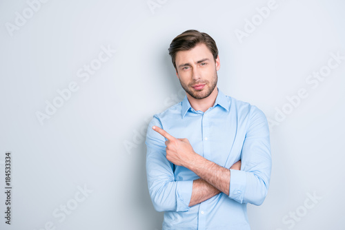 Advertisement concept. Portrait of cute, ideal, caucasian man in shirt, pointing and  to copy space with crossed arms, looking at camera, standing over grey background © deagreez