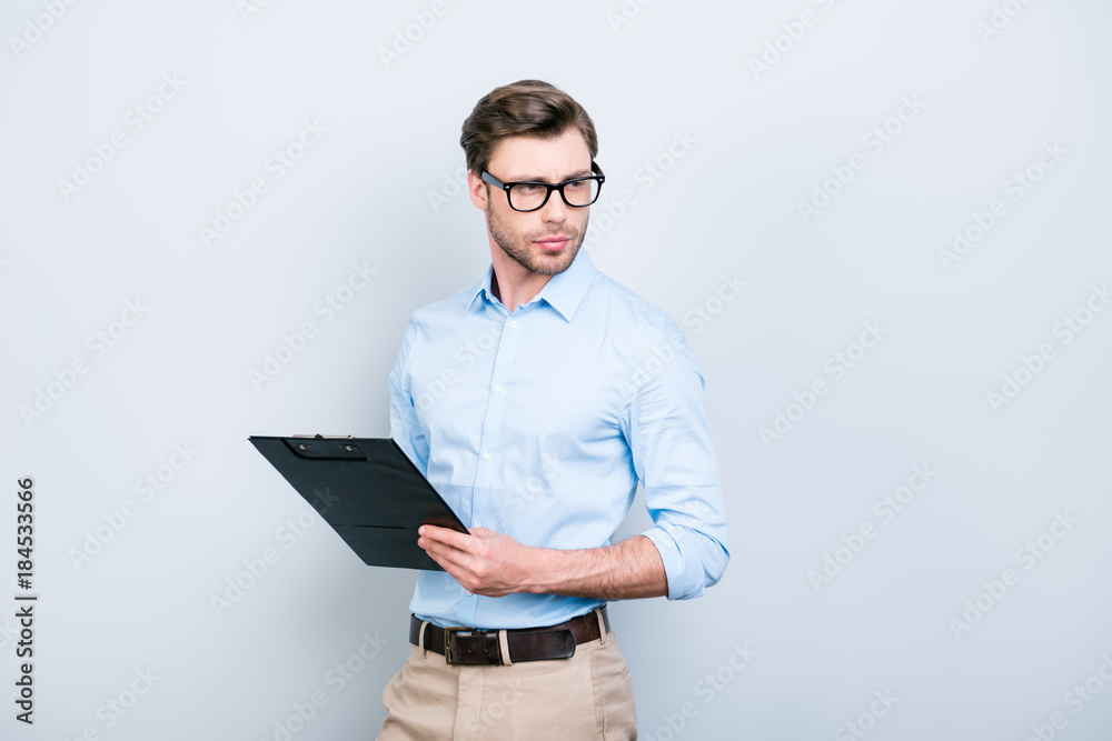 Close up portrait with copy space of attractive, clever, perfect man in shirt, pants, glasses holding clip board looking to the side over grey background Close up portrait with copy space of attractive, clever, perfect man in shirt, pants, glasses holding clip board looking to the side over grey background