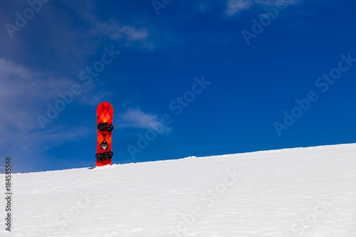 Distant view to red - orange board with black duck stance positioned bindings for snowboarding stuck straight into the snow with background of blue sky 