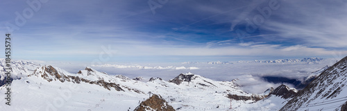 Panorama of high-altitude ski slopes and cable car lifts with foggy Alps mountains, blue sky and white clouds in background at glacier hills in March month