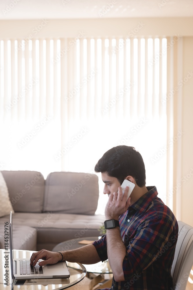 Man using laptop while talking on mobile phone in living room Man using laptop while talking on mobile phone in living room