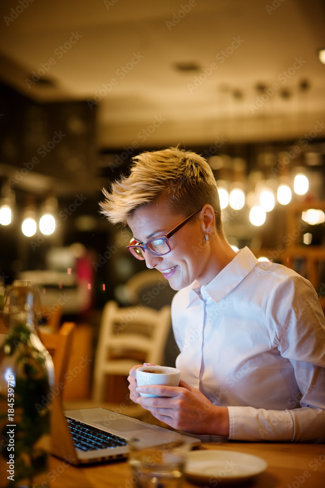 Close up side view of stylish confident smiling short hair blogger woman holding a cup of coffee while sitting in a cafe and working on a laptop at night. Close up side view of stylish confident smiling short hair blogger woman holding a cup of coffee while sitting in a cafe and working on a laptop at night.