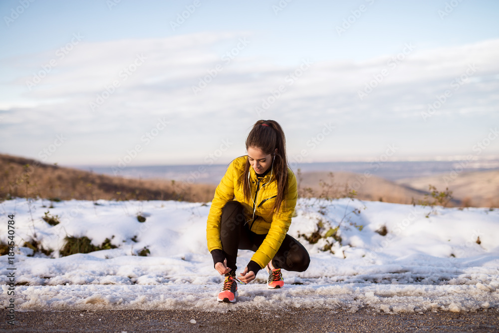 Young slim athletic girl tying shoelaces in winter sportswear on snowy winter road with earphones in the sunny morning. Young slim athletic girl tying shoelaces in winter sportswear on snowy winter road with earphones in the sunny morning.
