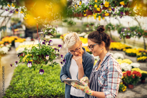 Adorable charming professional modern florist woman showing a list of flowers on a tablet to the curious attractive blonde female customer in a greenhouse.