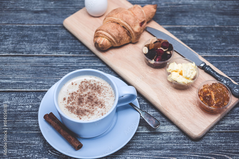 Croissant, orange jam, butter and coffee with milk and cinnamon. Continental breakfast concept. Wooden background