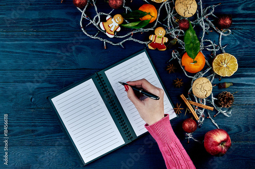 A girl with a red manicure in a knitted pink sweater writes a list of cases for the new year. A list of Christmas presents. Typical festive winter accessories, attributes, ornaments. photo