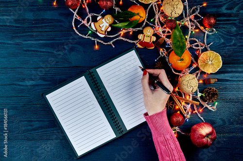 A girl with a red manicure in a knitted pink sweater writes a list of cases for the new year. A list of Christmas presents. Typical festive winter accessories, attributes, ornaments. photo