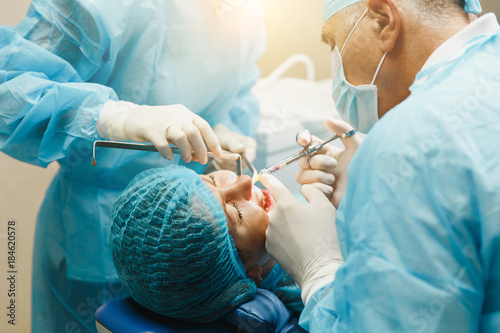 Male old professional dentist surgeon in uniform and female assistant helps to performing operation install dental implant teeth of woman patient in clinic light office with modern tools equipment. photo