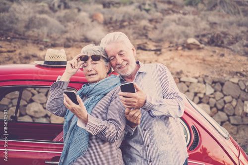 Elderly couple with hat, with glasses, with gray and white hair, with casual shirt, on vintage red car on vacation enjoying time and life. With a cheerful mobile phone smiling