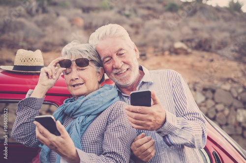 Elderly couple with hat, with glasses, with gray and white hair, with casual shirt, on vintage red car on vacation enjoying time and life. With a cheerful mobile phone smiling photo