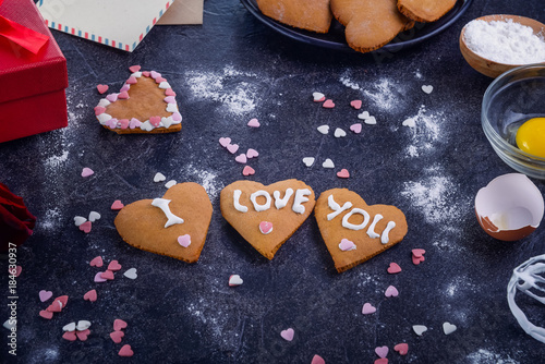 Homemade cookies in shape of heart with I Love you words as gift for lover on Valentine's day. Dark stone background with ingredients, flower and decor. Love gift concept. Selective focus. Copy space
