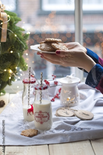 a girl drinks milk and eats cookies at a table by the window and a wreath of Christmas tree