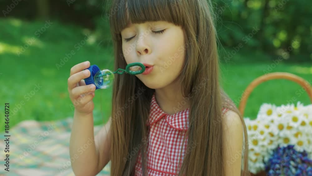Close up of the funny little girl with long hair blowing soap bubles while sitting in the nice green park. Summer period. Portrait. Outdoors