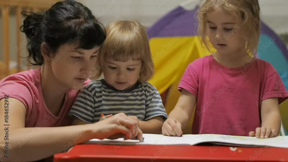 The educator and two little children girl and boy sitting at table drawing. Brother and sister and their mother drawing with colorful crayons and markers The educator and two little children girl and boy sitting at table drawing. Brother and sister and their mother drawing with colorful crayons and markers