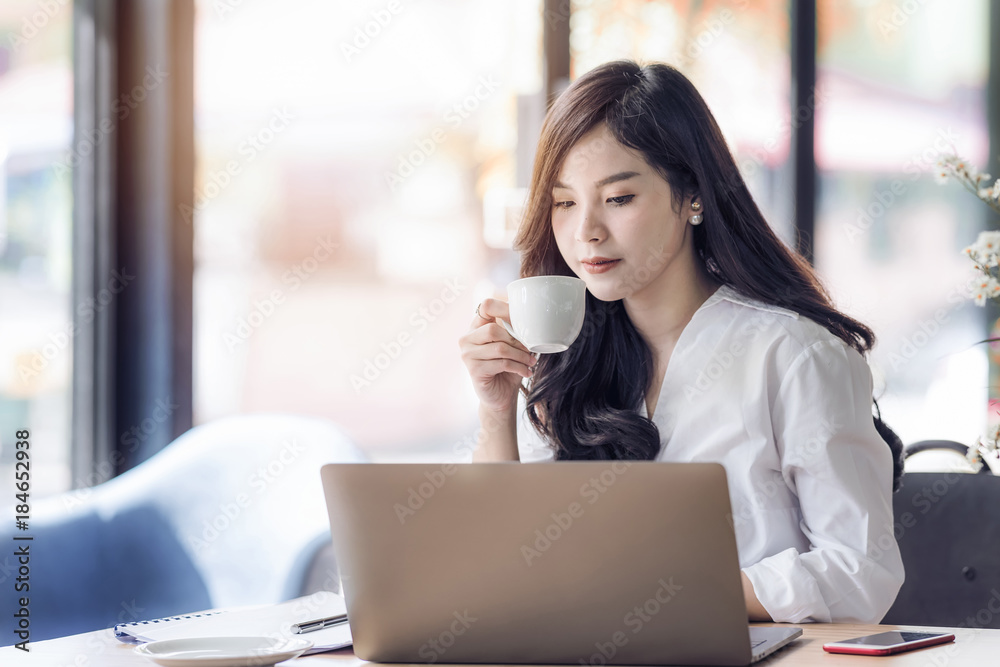 Young business asian woman holding cup of coffee and looking at laptop,woman officer  drink coffee at break time.