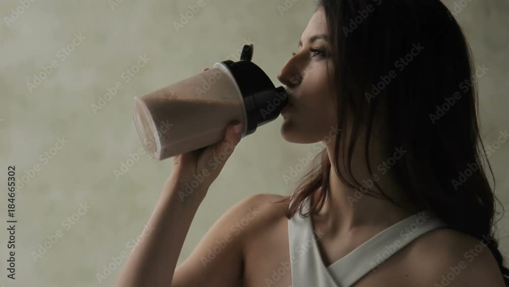 Attractive woman drinks protein cocktail is in apartment. Female opens plastic shaker, takes sip of wholesome beverage and smiles friendly, exposing teeth. Beautiful brunette dressed in trendy white