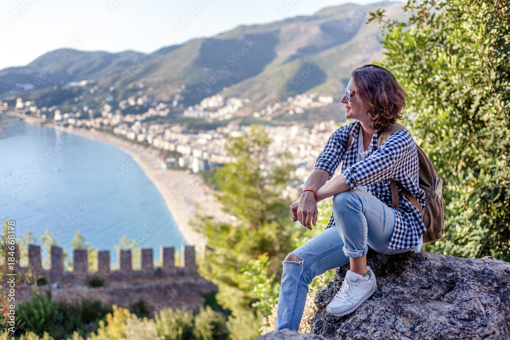 Naklejka premium young woman traveler with a backpack sitting and admiring the scenery with the sea and mountains at sunset